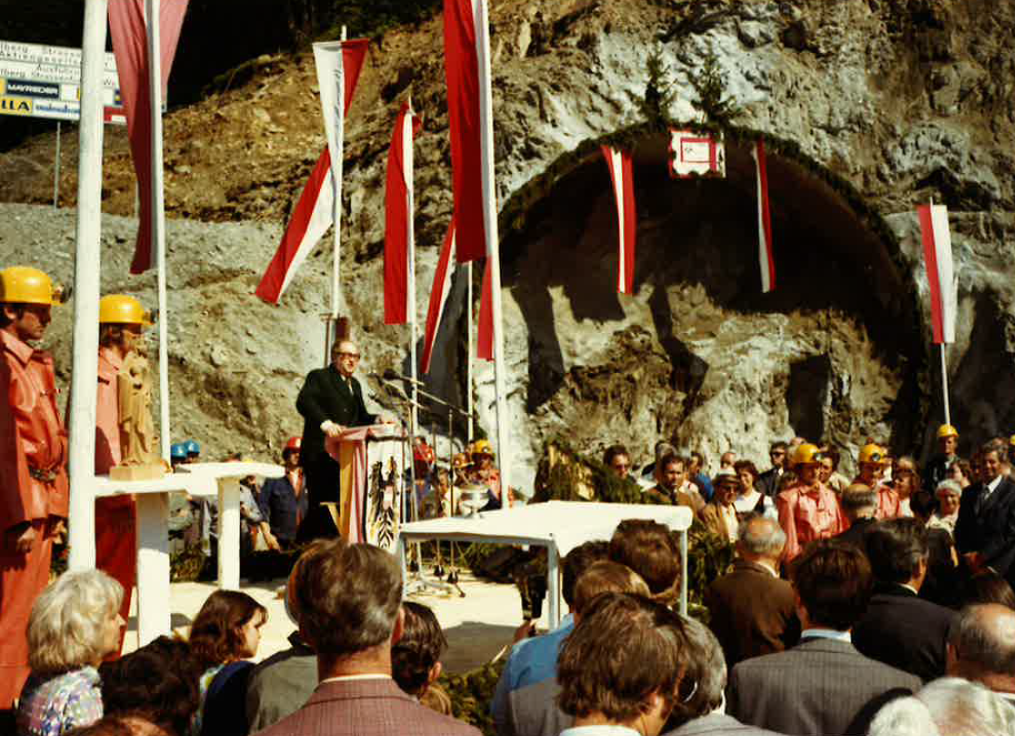 Eine Menschenmenge steht vor einem Podium, auf dem ein Mann vor einem Pult steht. Im Hintergrund ist das noch undurchbrochene Tunnelportal