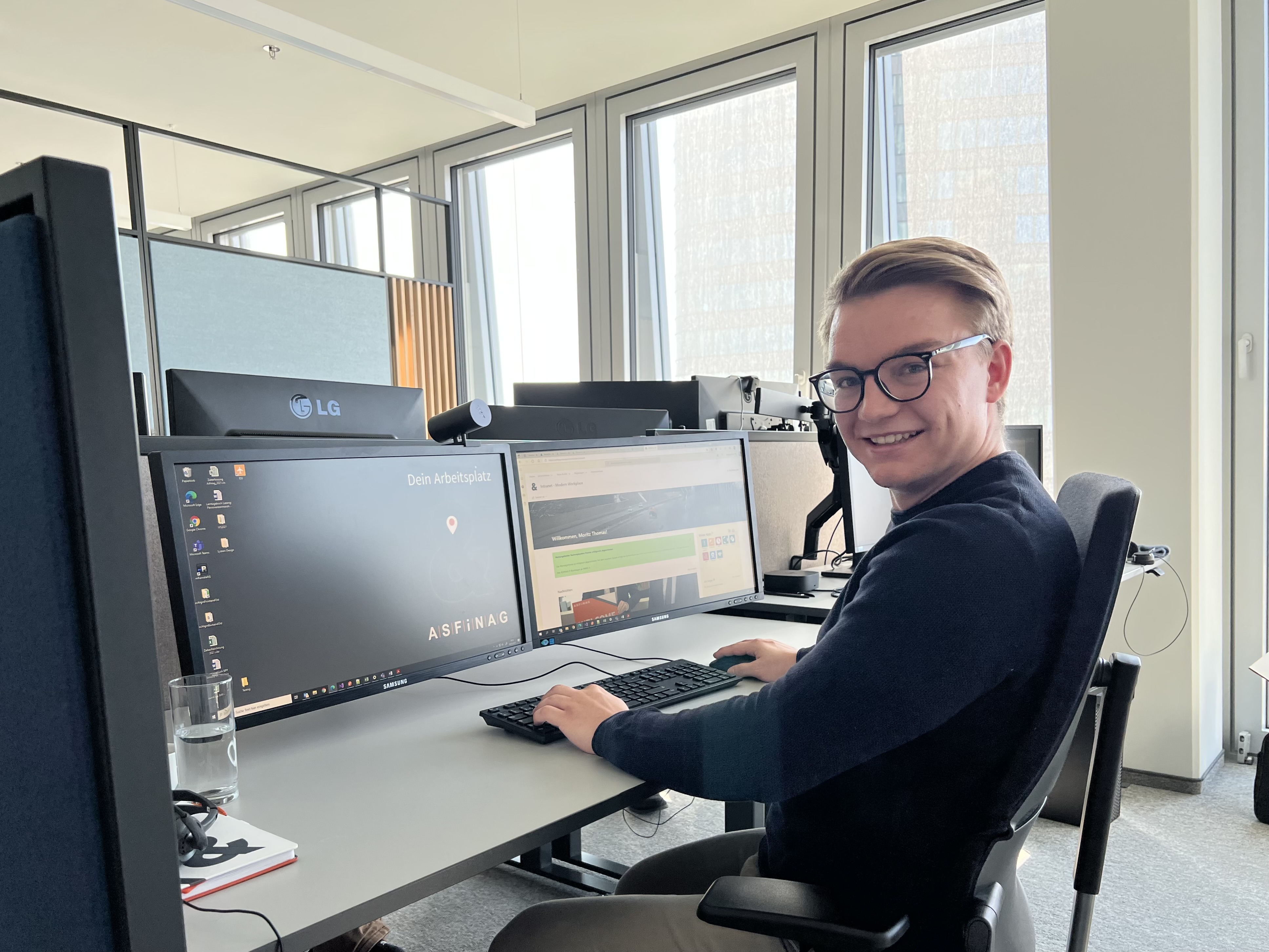 Ein junger Mann sitzt an einem Schreibtisch vor einem Computer und lächelt in die Kamera